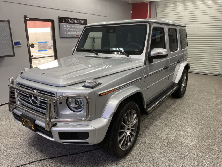 A silver Mercedes-Benz G-Class SUV is parked indoors on a speckled gray floor. The vehicle features distinctive headlights, a prominent front grille, and chrome accents. The background includes a closed garage door and a wall with a sign listing three combinations.