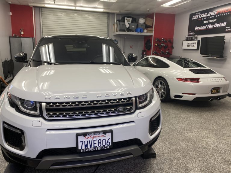 A white Range Rover and a white Porsche are parked inside a car detailing shop. The Range Rover is facing forward, while the Porsche faces towards the wall with shelves and tools. A sign reads "THE DETAILING PROS" and the floor is made of speckled gray material.