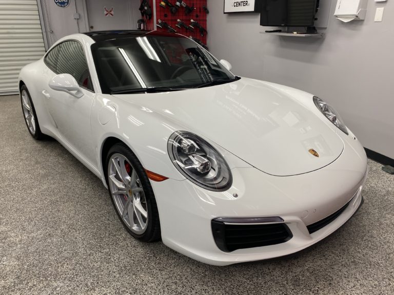 A white Porsche 911 sports car is parked indoors on a gray speckled floor. The car features a sleek design with a curved roof, shiny alloy wheels, and distinctive headlights. The right corner of the room displays various tools and equipment.