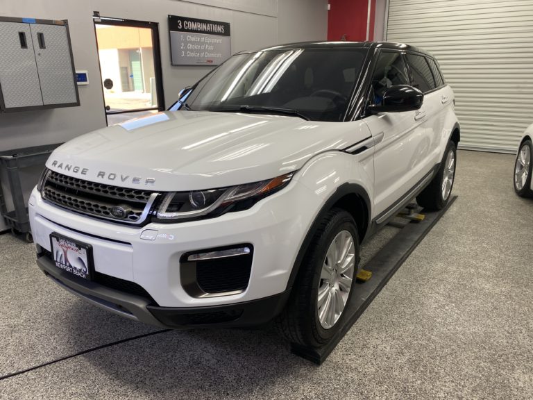 A white Range Rover SUV is parked indoors on a car lift. The vehicle is clean and shiny, and the background features a closed garage door and various workshop tools on the walls. The front license plate displays a design with a distinctive logo.