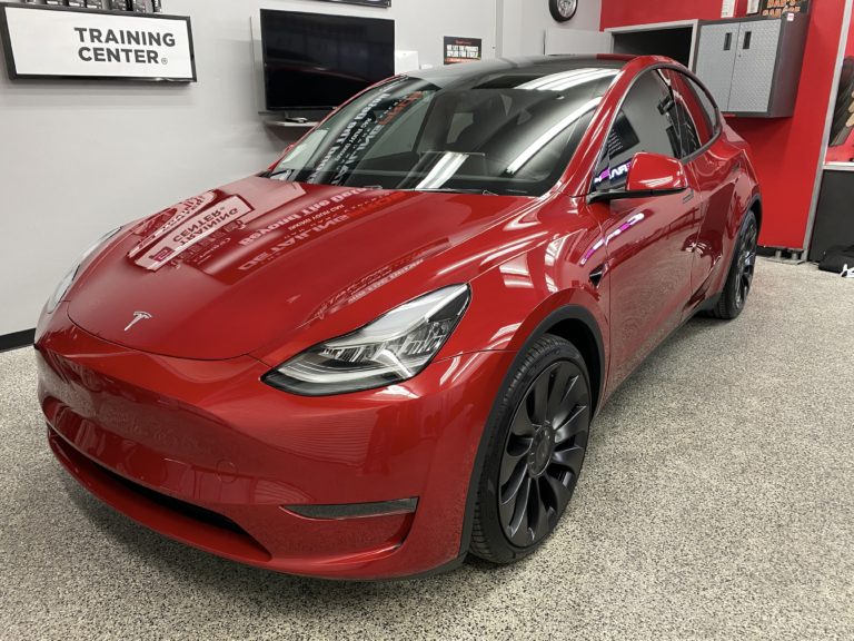 A shiny red Tesla electric car is displayed indoors in a showroom or training center. The vehicle features sleek, modern design elements and black wheels. The background includes a wall-mounted TV, a "Training Center" sign, and various car-related equipment.