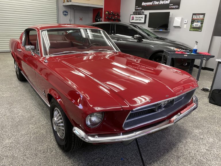 A shiny, red vintage Ford Mustang is parked in a well-lit garage with a polished floor. The car's sleek exterior is freshly detailed, and a black car is visible in the background. Various car care products and equipment are seen on a table to the right.