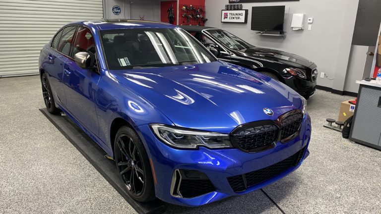 A blue BMW sedan is parked indoors on a gray, speckled floor. Next to it is a black car, partially visible. The background includes a roller door, a mounted TV, and shelves with various items. A sign that reads "Training Center" is visible on the wall.