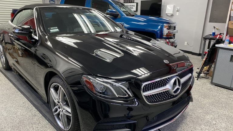 A sleek black Mercedes-Benz convertible is parked inside a garage. Behind it, a blue pickup truck is visible. The garage space has polished floors and is equipped with various tools and equipment. The car's front grille and headlights are prominently featured.