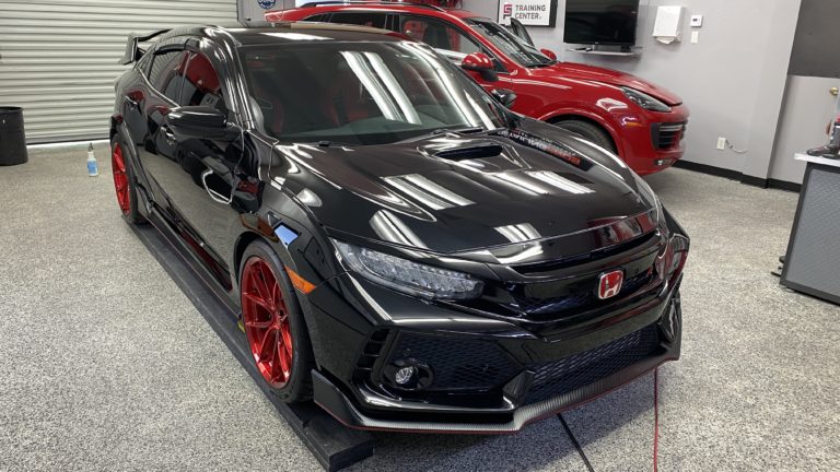 A black Honda Civic Type R with red accents and red custom wheels is parked inside a garage with overhead lighting. Another red SUV is partially visible in the background. The garage floor has a speckled finish, and a roller door is seen on the left.