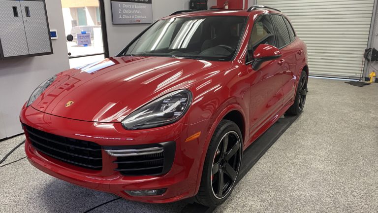 A red Porsche SUV is parked inside a garage with its front facing slightly to the left. It has black rims with a highlighted silver stripe. The garage walls are light-colored, and there is a large roll-up door in the background.