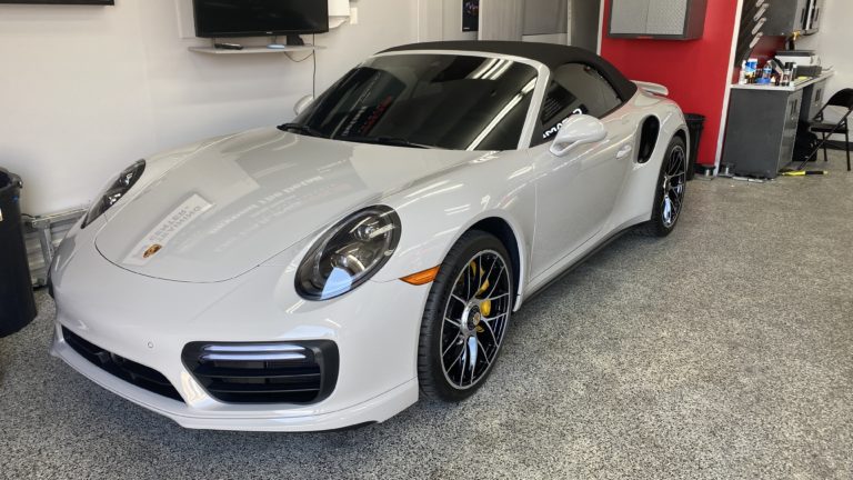 A sleek silver Porsche 911 convertible is parked inside a clean, modern garage. The car features a black soft top, black alloy wheels with yellow brake calipers, and reflective windows. The garage has a red and white interior, with tools and equipment visible in the background.