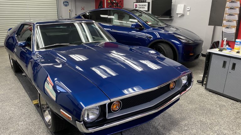 Two cars are parked inside a garage with a polished concrete floor. The car in the foreground is a vintage blue coupe with chrome details, while the vehicle in the background is a modern blue SUV. Shelves and tools are visible on the walls.