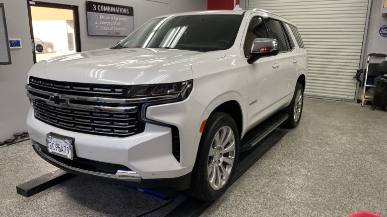 A white SUV with sleek design and chrome accents is parked inside a garage with a red wall. The vehicle is positioned on a lifting platform. The background includes a partially open roll-up door and various garage equipment. A sign lists "3 Combinations.