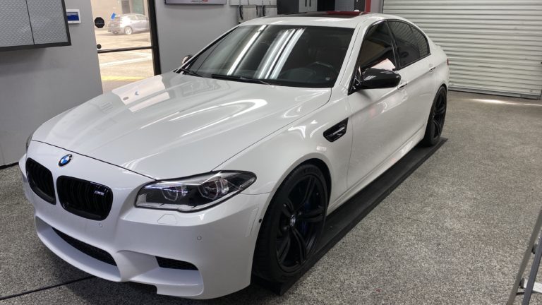 Image of a white BMW sedan parked inside a workshop. The car features a sporty design with black wheels, sleek headlights, and a black tinted window. The workshop includes a gray, speckled floor and various automotive equipment in the background.