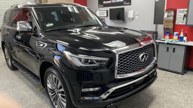 A glossy, black SUV is parked in a garage with various tools and supplies arranged on shelves and countertops. The car is clean and appears to be recently detailed. The wall has a sign that reads "Beyond the Detail.