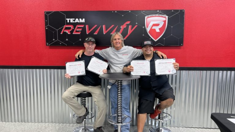 Three men pose with certificates while sitting and standing around a high table. They are in front of a red wall with a "Team Revivify" sign. All three appear happy and are holding their certificates proudly. The room has a modern industrial decor.