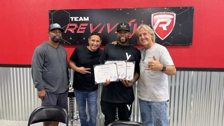 Four men are standing together in front of a "Team Revive" sign. The two men in the middle are holding certificates. All four are smiling and appear to be celebrating an achievement. The background includes a red wall with the Revive logo and a metallic lower wall section.