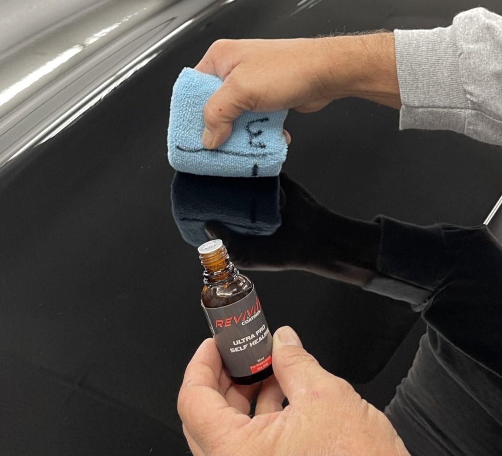 A person from Team REVIVIfy applies a liquid from a small bottle labeled "REVIVIFY" onto a black surface using a blue microfiber cloth. The person is wearing a gray long-sleeve top, and the black surface reflects parts of their hands, showcasing the precision in applying advanced coatings.