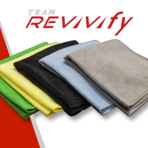 A variety of colored Revivify Microfiber Towels, including green, yellow, black, blue, and gray, are neatly stacked. The text "TEAM REVIVIFY" is displayed above the towels in a modern, italicized font. The background features a striking red and white diagonal color scheme.