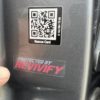 Close-up of a "Protected By Revivify" - Clear Vinyl 2" sticker on a surface, with the text "Protected by Revivify" and a finger pointing towards it. Above the sticker is a QR code labeled "Rescue Card," accompanied by some smaller alphanumeric characters.