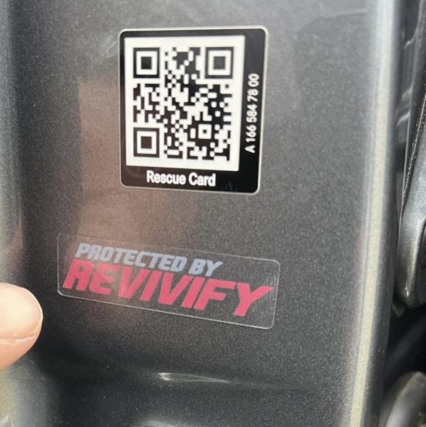 Close-up of a "Protected By Revivify" - Clear Vinyl 2" sticker on a surface, with the text "Protected by Revivify" and a finger pointing towards it. Above the sticker is a QR code labeled "Rescue Card," accompanied by some smaller alphanumeric characters.