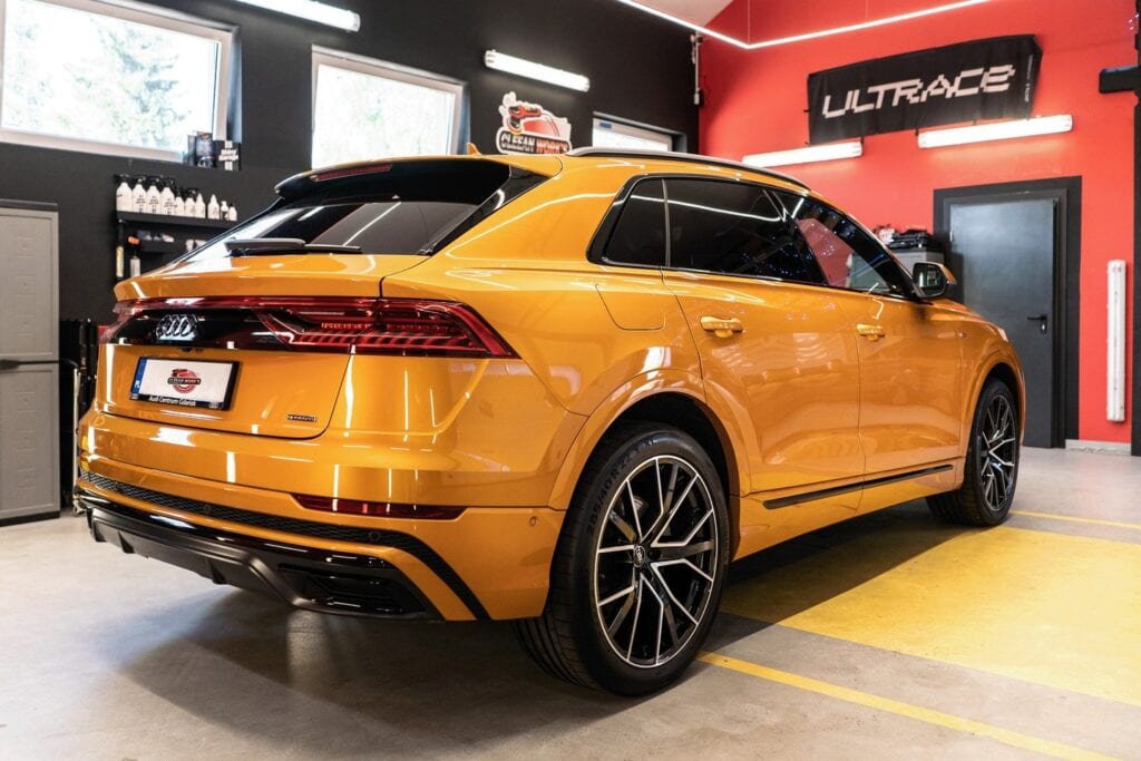 A sleek yellow Audi SUV parked inside a modern, well-lit automotive garage with a red and black color scheme. The car has dark tinted windows and black rims. The interior design includes shelves with car care products and a wall with the logo "ULTRACE" visible in the background.