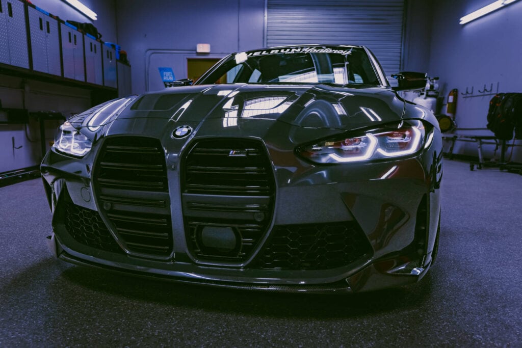 A sleek, black BMW car with illuminated headlights is parked in a dimly lit, modern garage. The car has a prominent front grille and reflective surface, showcasing its polished exterior. Shelves and racks are visible in the background.