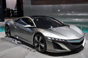 A sleek, silver sports car is showcased under bright lighting at an auto show. The car has a streamlined design with sharp angles, futuristic headlights, and large, distinctive black wheels. The logo on the front and "NSX Concept" written on the display platform are visible.