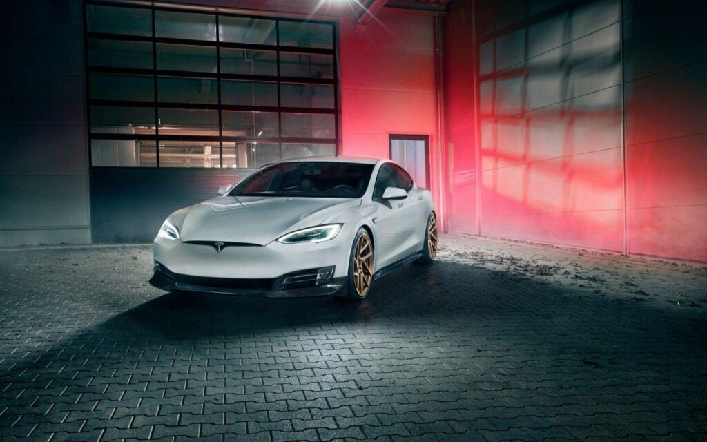 A silver Tesla Model S with custom bronze wheels is parked in an industrial area at night. The scene is illuminated with dramatic lighting, casting a red and blue glow on the surrounding walls. The car's headlights are on, emphasizing its sleek design.