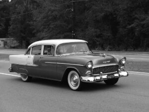 A black and white photo shows a vintage Chevrolet car driving on a road. The car has a two-tone paint job, with a lighter color on the roof and a darker color on the body. A driver is visible through the windshield, seemingly heading home as trees line the background.