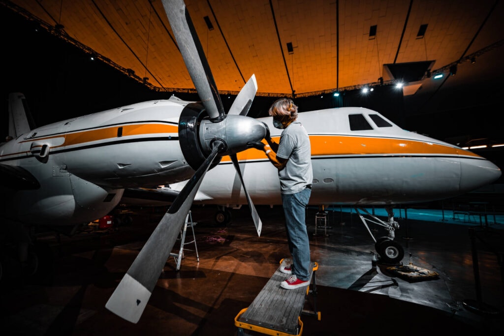 A person standing on a platform performs maintenance on the propeller of a white and yellow aircraft inside a hangar. Representing Team Revivify, they attend to an aircraft with multiple windows and two propellers, illuminated by the hangar's lights.