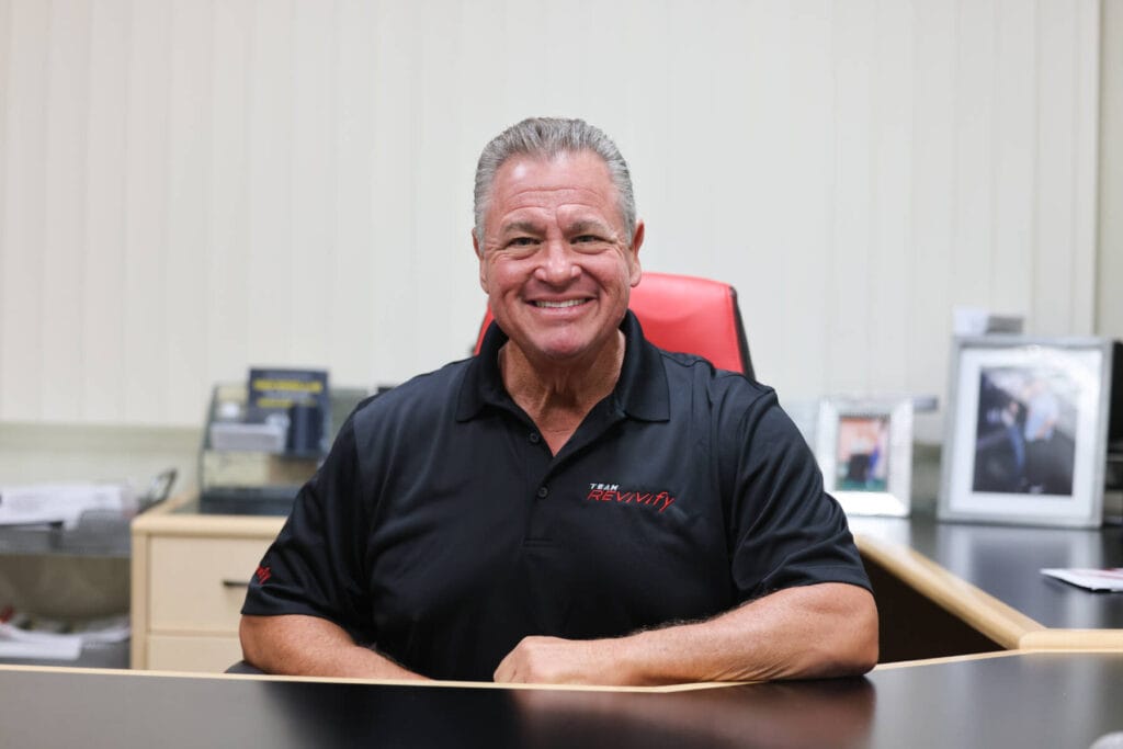 A man with grey hair and a warm smile sits at a desk in an office, exuding an air of support. He is wearing a black polo shirt with red and white text on it. There are documents, a curved monitor, and framed photographs on the desk and in the background.