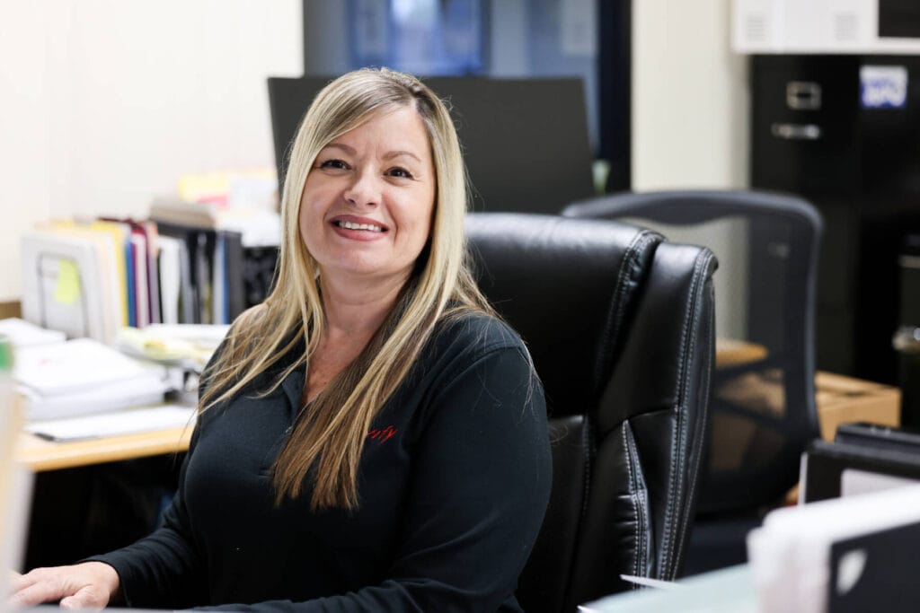 A woman with long blonde hair is sitting at a desk in an office, smiling at the camera. The desk has various papers and office supplies on it, emphasizing her organized approach to support tasks. Files and cabinets are in the background, and she is wearing a black long-sleeve shirt.