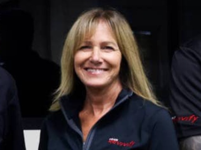 A woman with shoulder-length blonde hair is smiling. She is wearing a dark zip-up jacket with a small Team Revivify logo on the chest. The background is dark and indistinct.