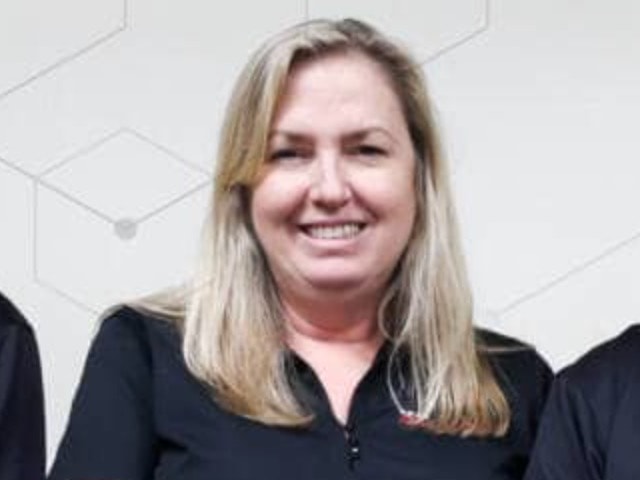 A smiling woman with shoulder-length blonde hair, wearing a black collared shirt, stands indoors against a light-colored wall with geometric patterns in the background, proudly representing Team Revivify.