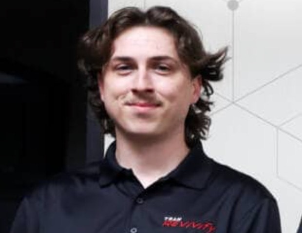 A person with shoulder-length dark hair and a mustache is smiling at the camera. They are wearing a black collared shirt with a red logo on the left side that appears to read "Team Revivify." The background features a dark area and a light section with geometric lines.