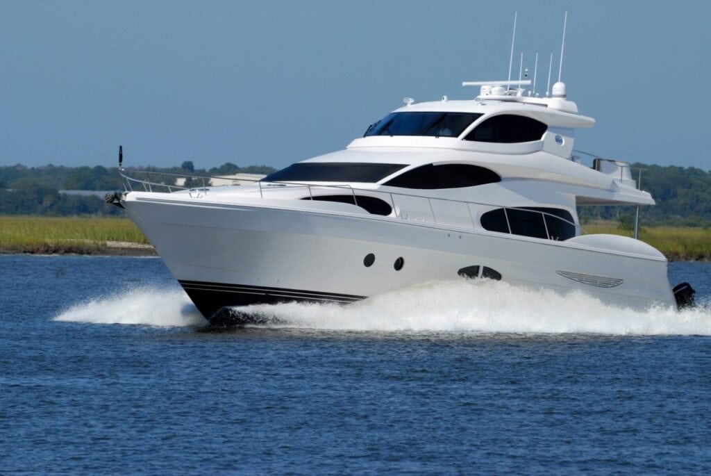 A white luxury yacht, coated in Revivify Coatings Marine for ultimate protection, glides through calm blue waters, creating a wake behind it. The background features a low, green shoreline under a clear blue sky.