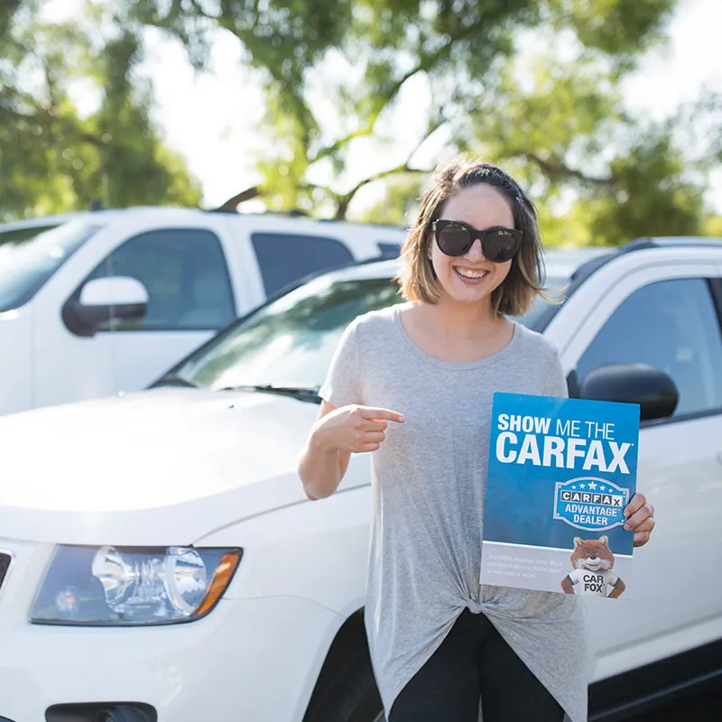 A woman wearing sunglasses and a gray shirt stands in front of a white SUV in a parking lot. She is smiling and pointing to a "Show Me the Carfax" report that she is holding, showcasing the CARFAX Partnership as key to boosting vehicle resale value.