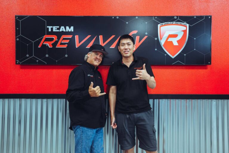 Two people stand in front of a black and red sign that reads "Team Revivv." They are both smiling and making a hand gesture with their thumbs and pinky fingers extended. One person is wearing a black hat and shirt, while the other is in a black polo and shorts, likely posing for the Revivify Coatings Gallery.