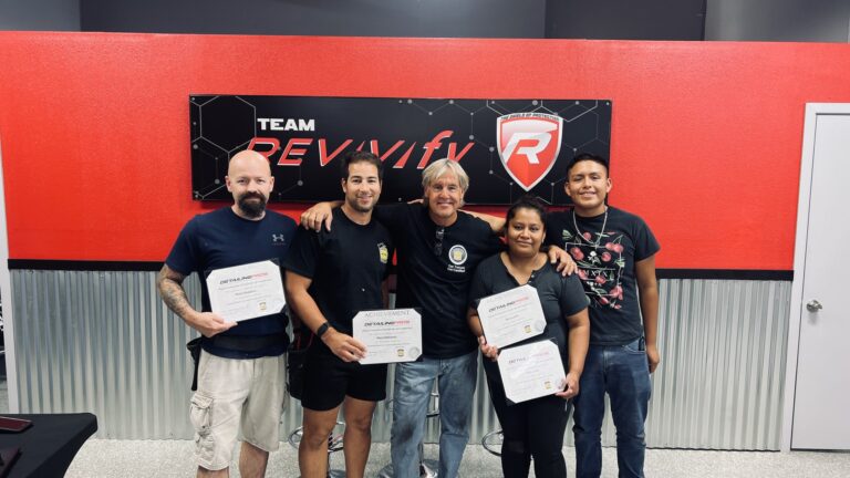 A group of five people stand together smiling and holding certificates in front of a red and black wall with a logo that reads "Team Revivify." They appear to be celebrating an achievement at the Revivify Coatings Gallery.