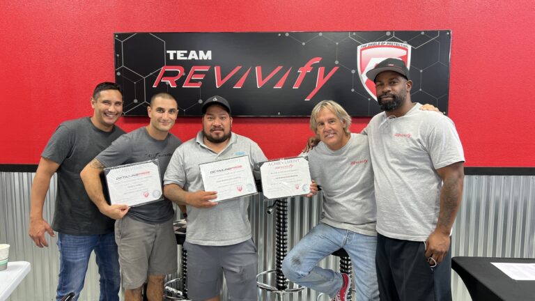 Five men are standing and posing for a photo in front of a red and black "Team Revivify" sign at the Revivify Coatings Gallery. Four of the men are holding certificates and all are dressed casually in t-shirts. They seem happy and proud, smiling broadly at the camera.