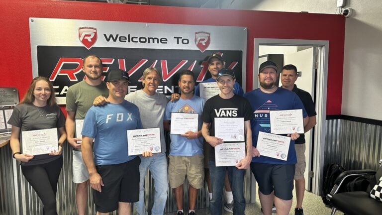 A group of nine people, eight men and one woman, stand in front of a "Welcome To REV'V12" sign inside the Revivify Coatings Gallery. They are holding certificates and smiling, suggesting an achievement or graduation event. The background features red and grey walls.