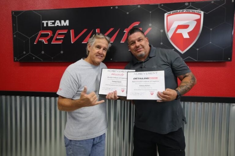 Two men standing in front of a "Team Reviva" backdrop at the Revivify Coatings Gallery, smiling and holding certificates of achievement. The man on the left is making a "shaka" hand gesture. Both are dressed casually, one in a grey shirt and the other in a dark shirt.