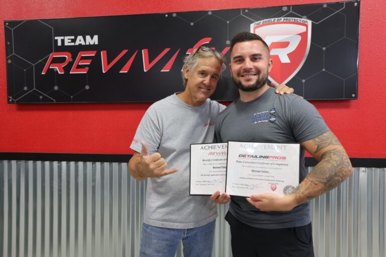 Two men are standing in front of a Team Revivif backdrop. The younger man on the right is holding a certificate and smiling at the camera, while the older man on the left gestures with a hand sign and also smiles. Both are dressed in casual shirts, celebrating their moment at the Revivify Coatings Gallery event.