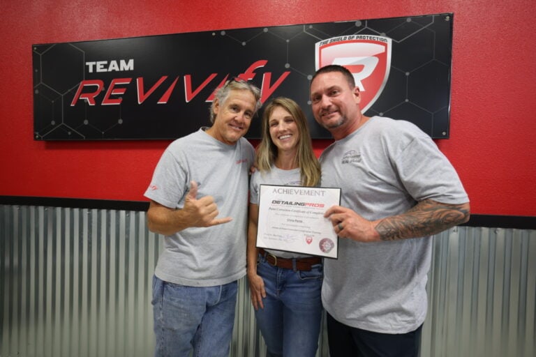 Three people stand smiling in front of a red and black "Team Revivify" sign. The person in the middle holds an Achievement certificate while being embraced by the other two. They are all wearing light gray shirts, celebrating their moment at the Revivify Coatings Gallery.