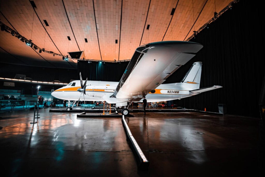 A polished twin-engine aircraft, featuring the latest Revivify Coatings, is displayed under dramatic lighting in an indoor setting with a high ceiling. Mostly white with yellow and black accents, it stands on a pristine, reflective floor. Multiple people and equipment are visible around the aviation marvel.