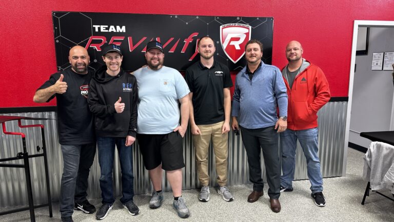 Six men standing in front of a "TEAM REVivf" sign on a black and red wall, posing for a photo at the Revivify Coatings Gallery. Four men are wearing casual clothing, and two are in business casual attire. One man on the left is giving a thumbs-up, and they all seem to be smiling.