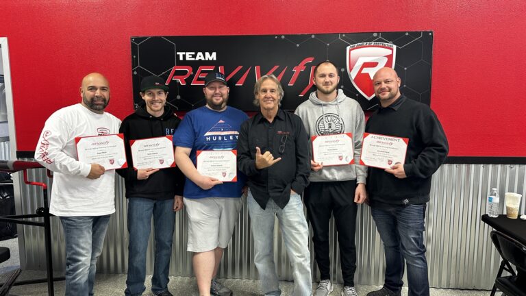 Six men stand side by side holding certificates and smiling in front of a 'Team Revive' sign on a red and black wall at the Revivify Coatings Gallery. They appear to be celebrating an achievement or milestone. One man in the center gestures with a "shaka" hand sign.