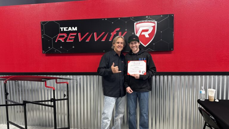 Two people stand together in front of a "Team Revivify" sign at the Revivify Coatings Gallery, with one person giving a thumbs up and the other holding a certificate. They are smiling and standing in a room with a red and black color scheme and corrugated metal paneling along the lower wall.