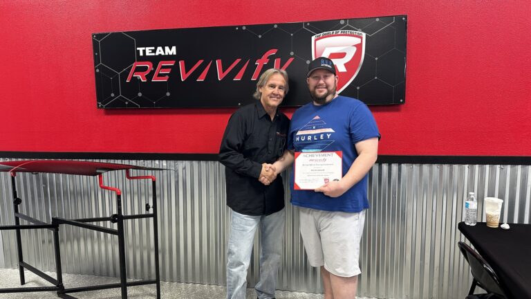 Two men shaking hands in front of a red and black wall with the text "TEAM REVIVify" on it. One man wears a black shirt, gray pants and long hair, while the other is dressed in a blue T-shirt and cap, holding a certificate with both hands. Welcome to the Revivify Coatings Gallery.