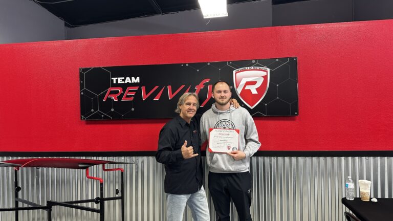 Two men standing in front of a red and black wall with a "Team Revivify" sign. The man on the left is making a thumbs-up gesture, while the man on the right holds a certificate of achievement. Both are smiling brightly. A nearby table, part of the Revivify Coatings Gallery, has cups and a water bottle visible in the foreground.