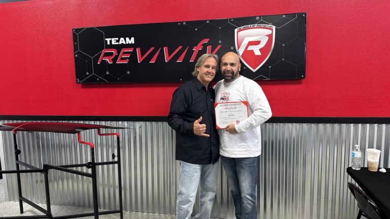 Two men are standing in front of a red and black "Team Revivify" sign. The man on the left is wearing a black shirt and making a thumbs-up gesture. The man on the right, holding an "Achievement" certificate and wearing a white shirt, smiles at the camera in this vibrant Revivify Coatings Gallery scene.